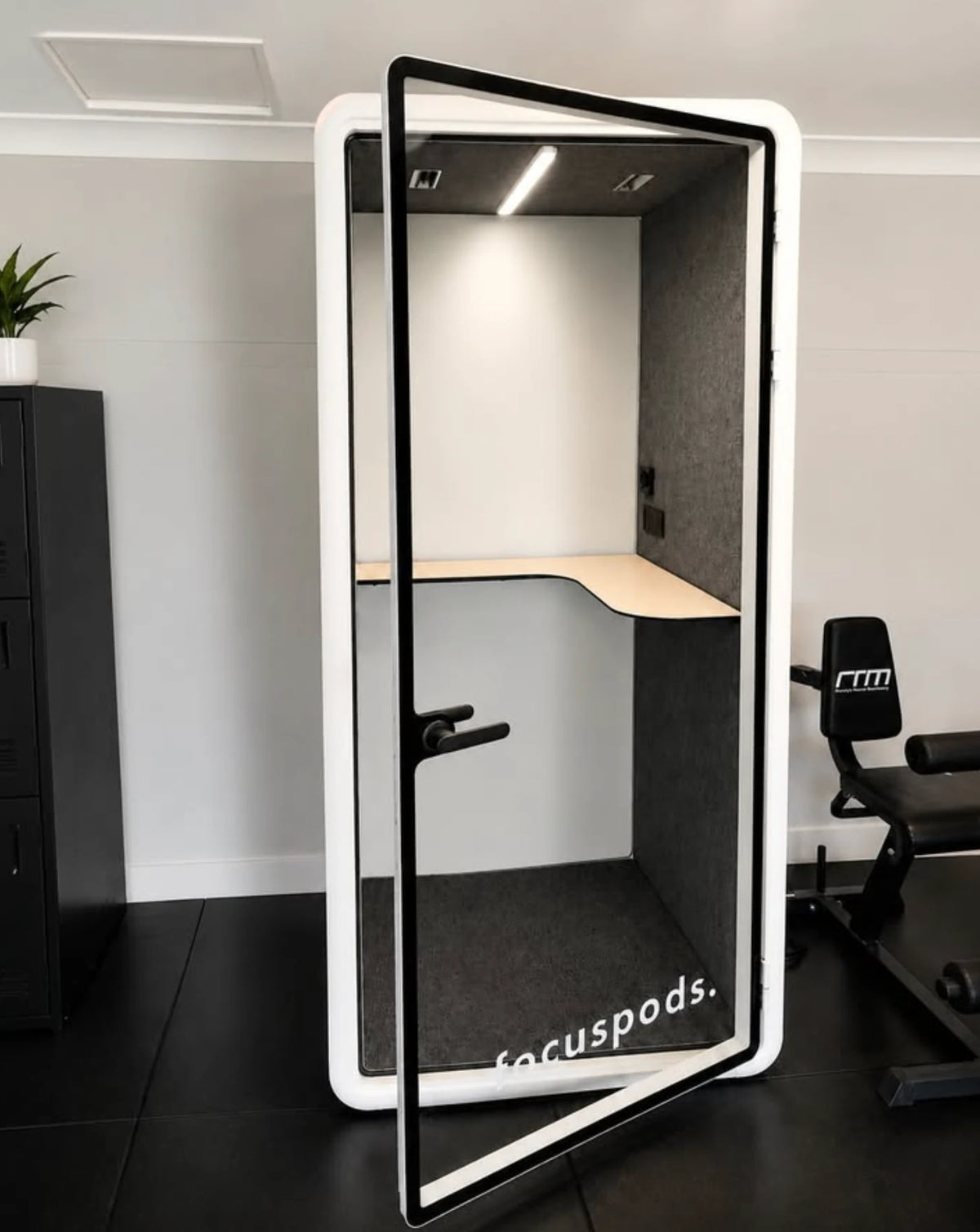 White FocusPod Solo with an open glass door and desk shelf in a Newcastle garage, beside home gym equipment.